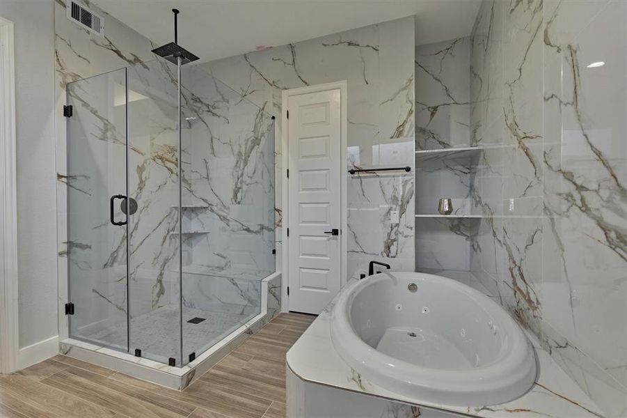 Stylized focal wall in the primary bath covered in tile for a unified look. Rain shower head and seat in the seamless glass shower. Built in shelves at the jetted tub and in the shower provide extra storage/display.