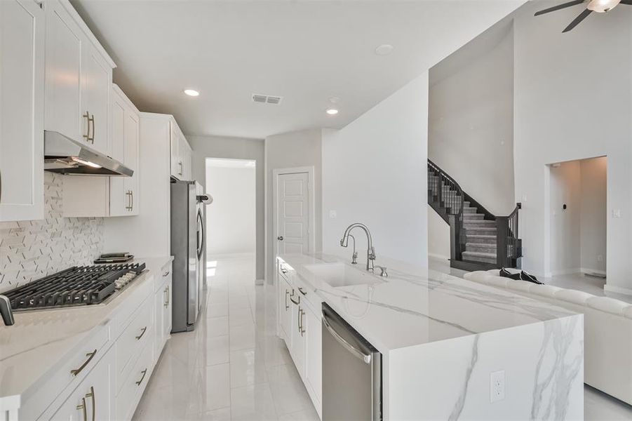 This is a modern kitchen with white cabinetry and marble countertops, featuring stainless steel appliances and a herringbone tile backsplash. It opens to a spacious area with high ceilings and a grand staircase, providing a bright and airy feel.