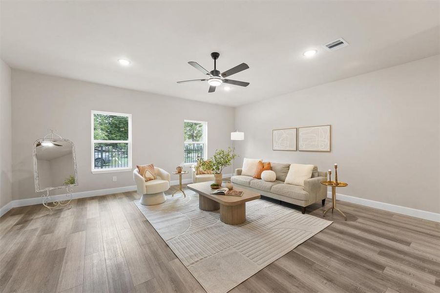 This is a spacious, modern living room featuring wood flooring, large windows with a view of a fence, neutral color tones, and contemporary furnishings. It has a ceiling fan and recessed lighting.