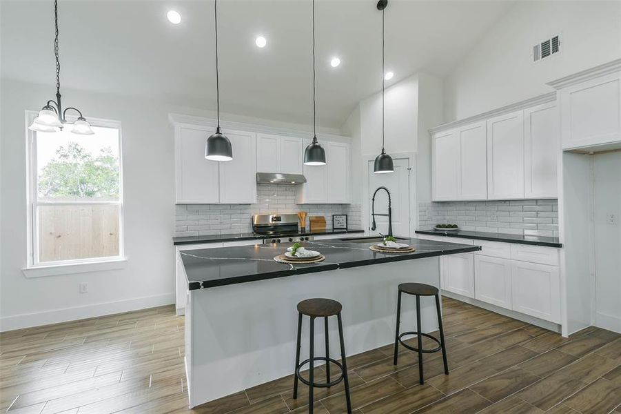 This is a modern kitchen with white cabinetry, stainless steel appliances, dark countertops, and a subway tile backsplash. It features an island with bar seating and pendant lighting, set against hardwood floors.