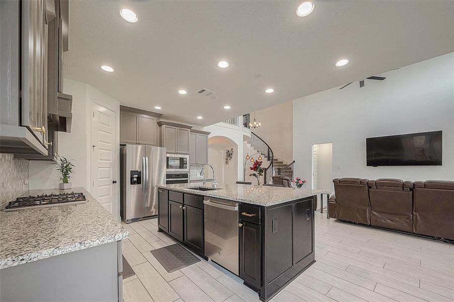 Modern open-concept kitchen with stainless steel appliances, granite countertops, and an island, flowing seamlessly into a cozy living area with ample seating and a flat-screen TV. The space is well-lit and features contemporary finishes.