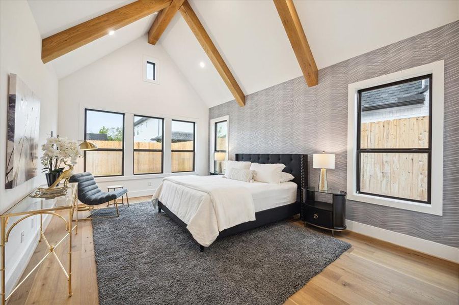 The serene primary bedroom features vaulted ceilings with decorative wood beams, wood floors,designer wallpaper, a transitional chandelier and views onto the back yard.