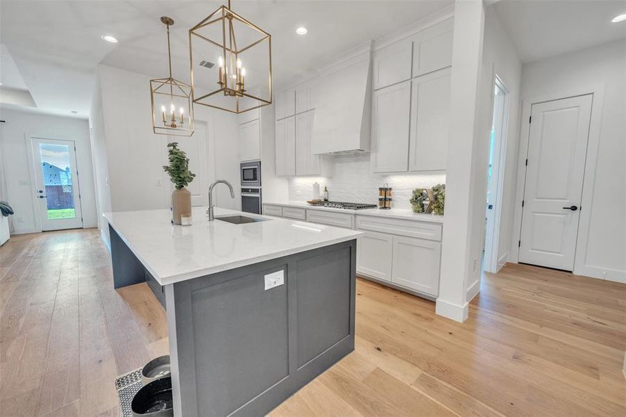 Kitchen featuring white cabinets, light wood-type flooring, hanging light fixtures, and an island with sink