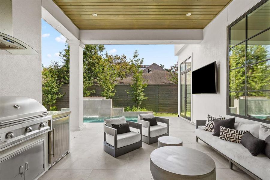 This inviting covered patio features a sleek wood-paneled ceiling with built-in recessed lighting and a ceiling fan for optimal comfort while expansive black-framed glass doors and windows provide a seamless connection between the indoor and outdoor living spaces.