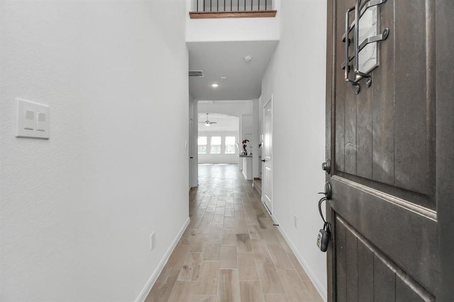 This is an interior view of a hallway leading into a bright, open-concept space. The hallway features light flooring and neutral wall colors, with a high ceiling that contributes to a spacious feel. A modern-styled door on the right adds a touch of elegance to the entrance.