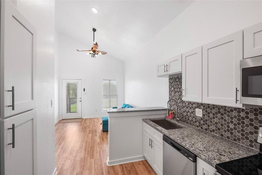 The is a similar view to the front entrance from the rear of the kitchen, showing cabinetry, granite, backsplash, ceiling fan, and the glassed front door with functional internal blinds.