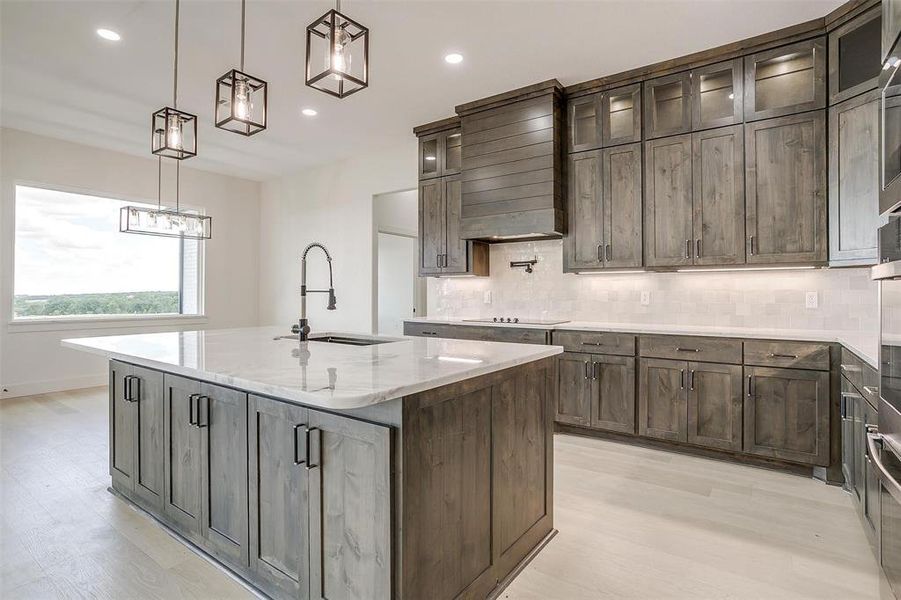 Kitchen featuring pendant lighting, custom exhaust hood, light stone counters, an island with sink, and sink