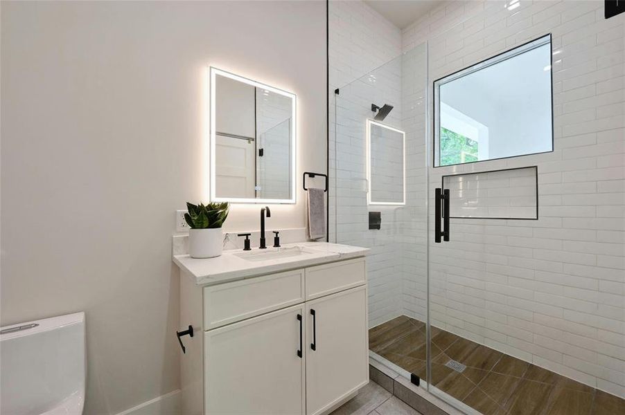 The modern bath on the second floor complements the home's aesthetic with white marble countertops, creating a seamless design theme throughout the home. The shower area is accentuated with wood tile, adding a natural element and a touch of warmth to the sleek and sophisticated bathroom design.