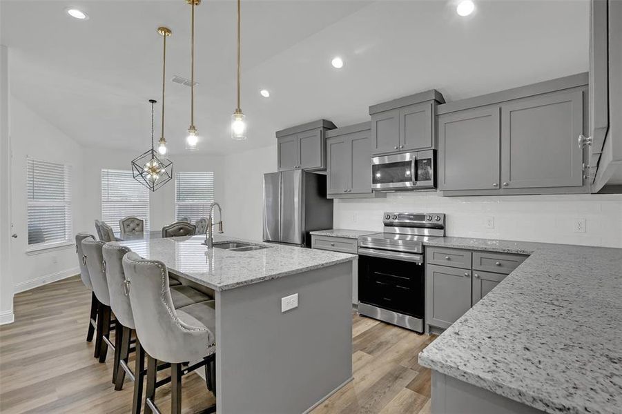 Kitchen with sink, appliances with stainless steel finishes, light stone countertops, decorative light fixtures, and an island with sink