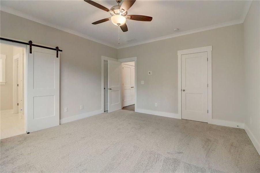 Unfurnished bedroom with crown molding, a barn door, light colored carpet, ensuite bathroom, and ceiling fan