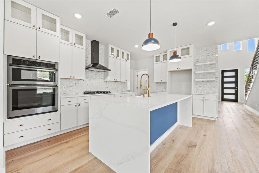 Kitchen with light hardwood / wood-style flooring, wall chimney range hood, backsplash, a kitchen island with sink, and appliances with stainless steel finishes