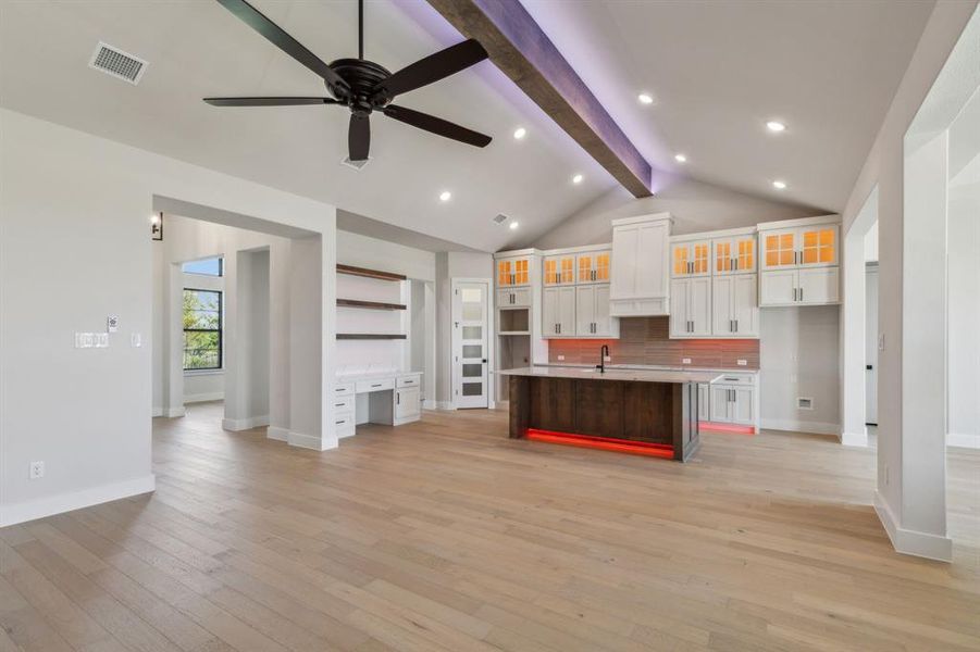 Kitchen featuring light wood-type flooring, a center island with sink, built in desk, white cabinets, and vaulted ceiling with beams