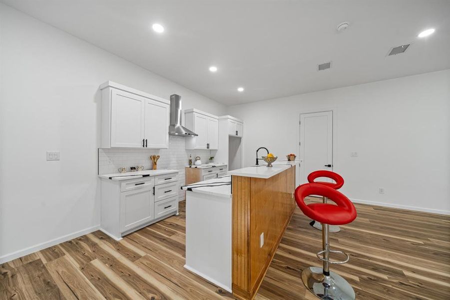Kitchen featuring tasteful backsplash, white cabinets, hardwood / wood-style floors, wall chimney range hood, and an island with sink