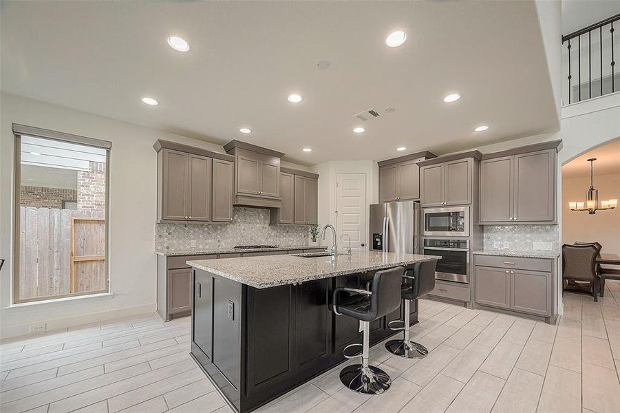 This is a modern kitchen with stainless steel appliances, granite countertops, a central island with bar seating, and ample cabinetry. There's a view to the backyard through a window and an adjoining dining area visible in the background.