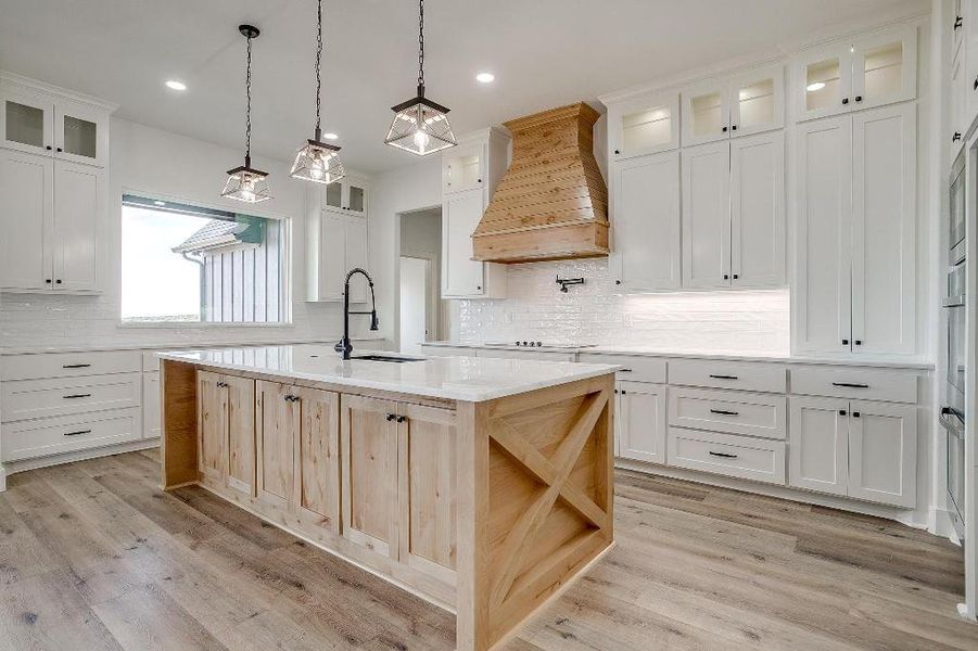 Kitchen with backsplash, custom exhaust hood, light hardwood / wood-style floors, and a center island with sink