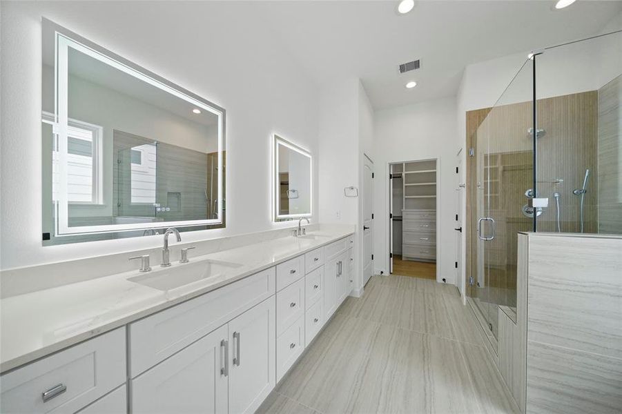 Modern bathroom featuring dual sinks with a large mirror, ample cabinet space, a glass-enclosed shower, and direct access to a walk-in closet. Bright and airy with a clean, contemporary design.
