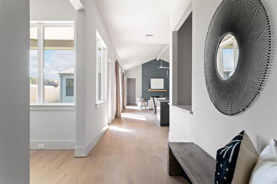 This is a modern hallway featuring natural light from large windows, sleek hardwood floors, neutral color walls with a contrasting accent wall, and contemporary decor elements like a unique mirror. A glimpse into the adjoining room suggests an open-plan layout.