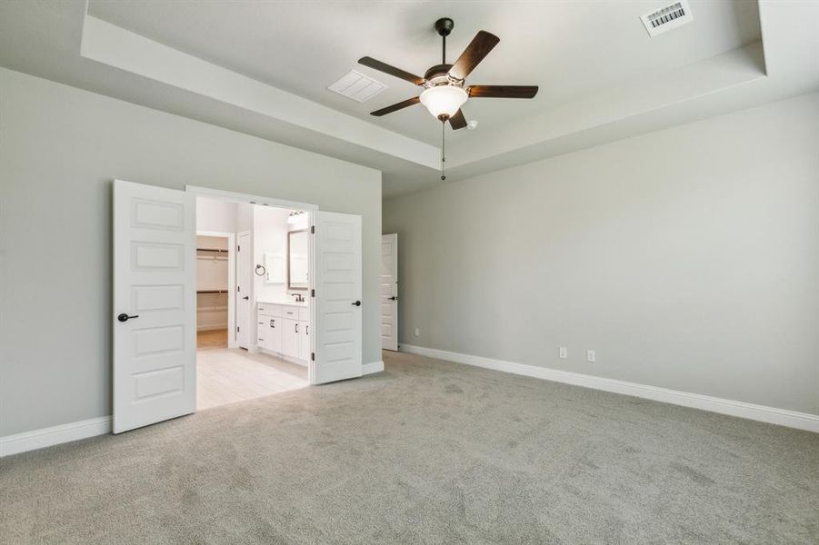 Unfurnished bedroom with connected bathroom, ceiling fan, a tray ceiling, and light colored carpet