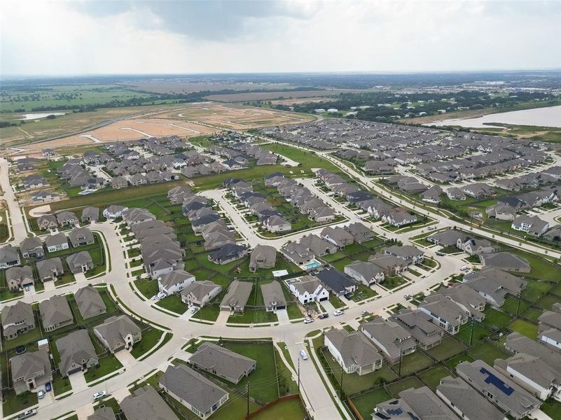 Aerial view of a sprawling suburban neighborhood with uniform houses and well-maintained lawns, featuring curving streets and cul-de-sacs, with open land and a large body of water nearby, suggesting room for community growth and recreational opportunities.