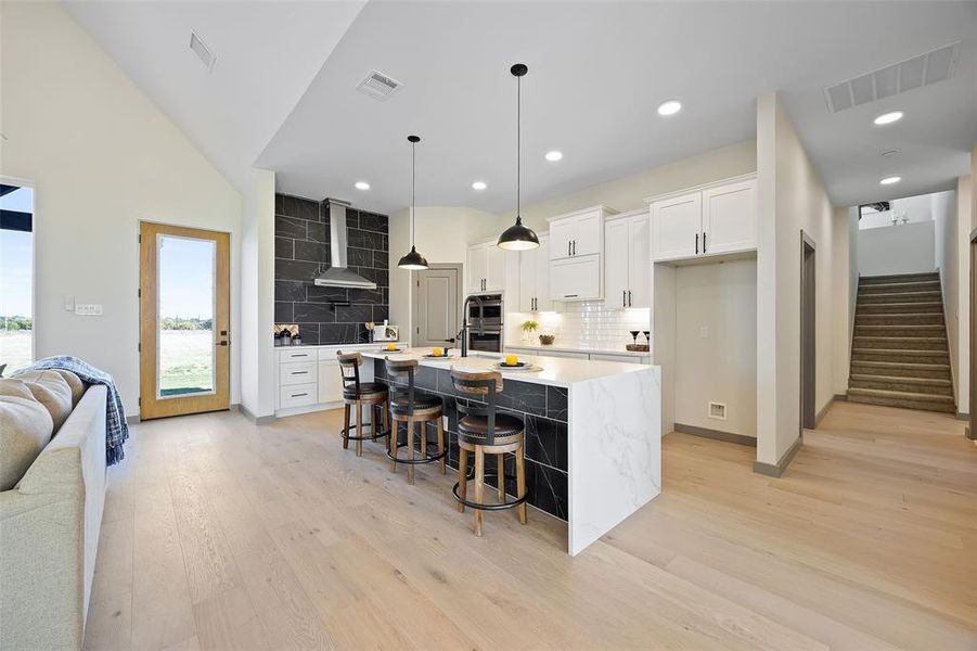 Kitchen featuring stainless steel double oven, a kitchen island with sink, wall chimney range hood, white cabinetry, and light hardwood / wood-style floors