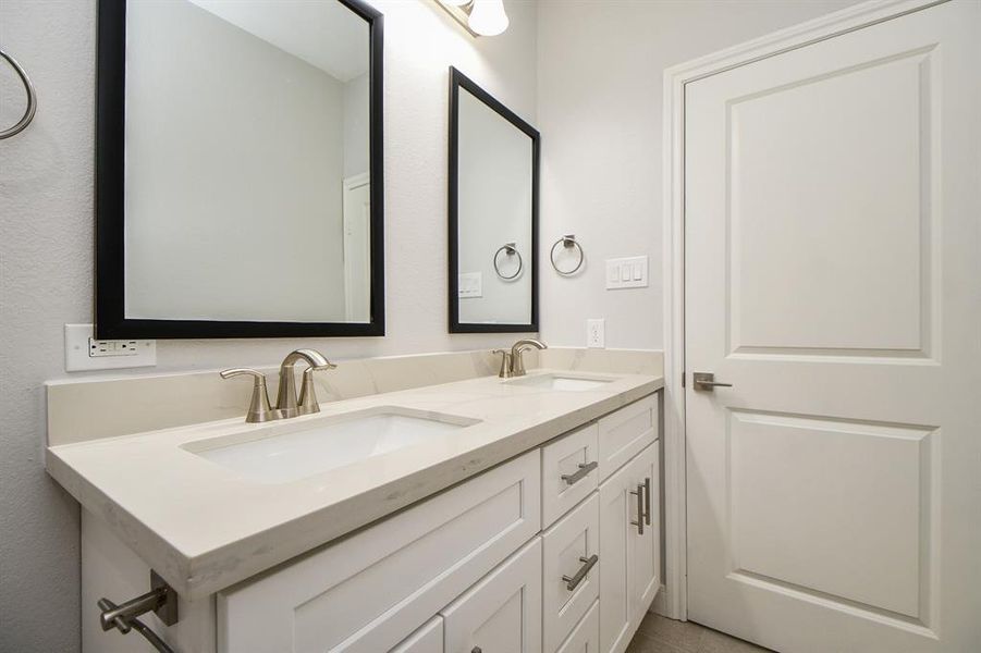 This is a modern secondary bathroom featuring a double vanity with a marble countertop, undermount sinks, and brushed nickel faucets. There are two framed mirrors above the vanity, and the space is completed with clean, white cabinetry offering ample storage. A closed door suggests additional privacy or access to another area.