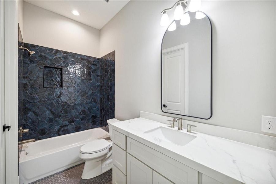 This modern bathroom features a sleek white vanity with marble countertop, a large mirror, and a bathtub with dark, hexagonal tile surround. It has a clean, contemporary design with bright lighting.