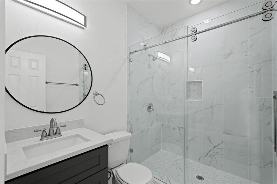 A large walk-in shower with modern tile detailing and recessed lighting, granite countertops, a framed mirror, and wooden cabinetry.