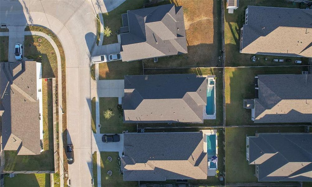 This aerial photo shows a residential neighborhood with single-family homes, each with a distinct backyard, some featuring pools. The streets are laid out in a curved pattern with driveways and cars, indicating a suburban setting.