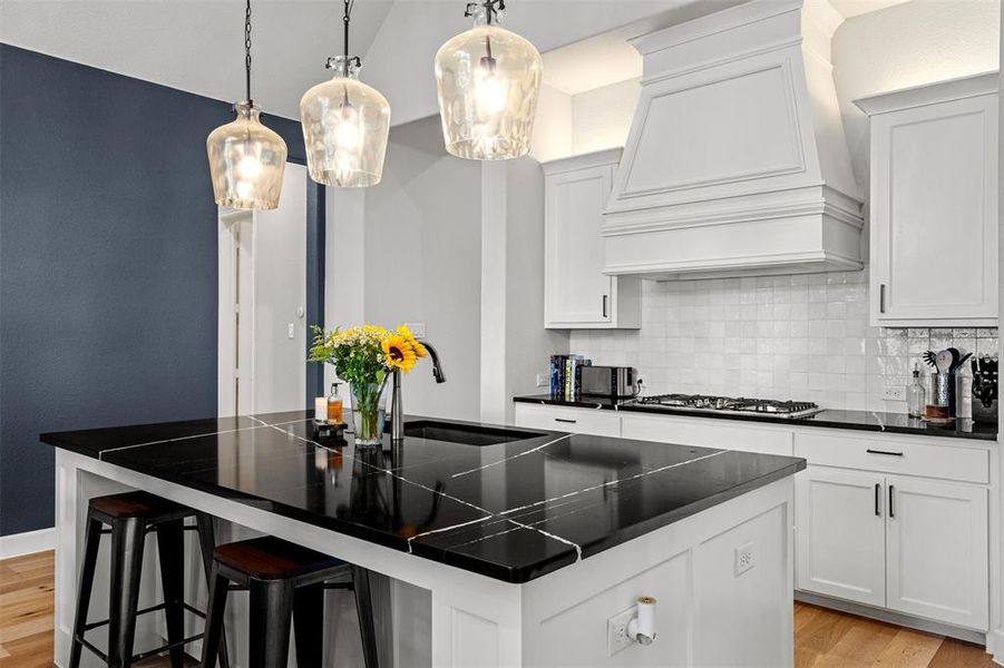 Kitchen featuring light wood-type flooring, a center island with sink, custom range hood, sink, and tasteful backsplash