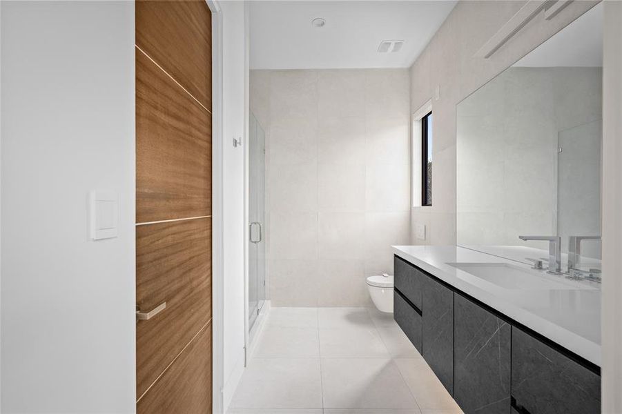 This sleek secondary bathroom features a minimalist design with large-scale neutral tile walls that give the space a modern and airy feel. The spacious vanity is adorned with black marbled cabinetry with subtle accent lighting and a white quartz countertop. The frameless glass shower enclosure complements the clean lines and openness of the space.