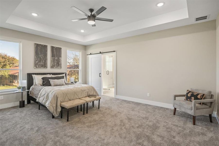 Carpeted bedroom with a barn door, ceiling fan, a tray ceiling, and connected bathroom