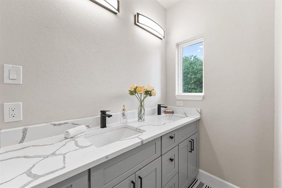 This is a modern bathroom featuring a double vanity with marble countertop, undermount sinks, and black fixtures. There's ample storage with gray cabinetry, and the room is brightened by natural light from a window and sleek light fixtures above.