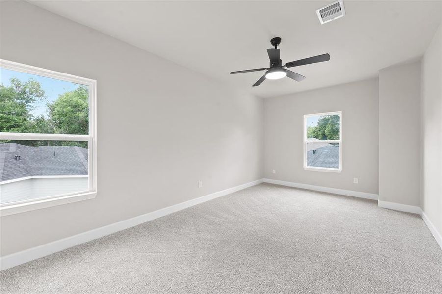 This is a bright, empty room featuring a fresh, neutral color palette, with plush carpeting, two large windows providing natural light, and a modern ceiling fan for comfort.