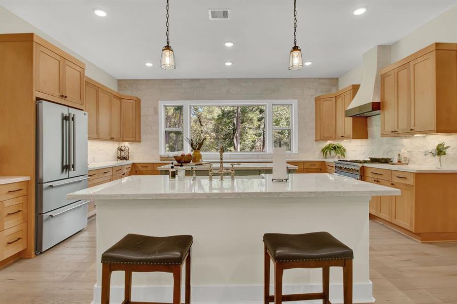 This is a spacious, modern kitchen with natural wood cabinets, stainless steel appliances, a large central island with seating, and a bright view of the outdoors.