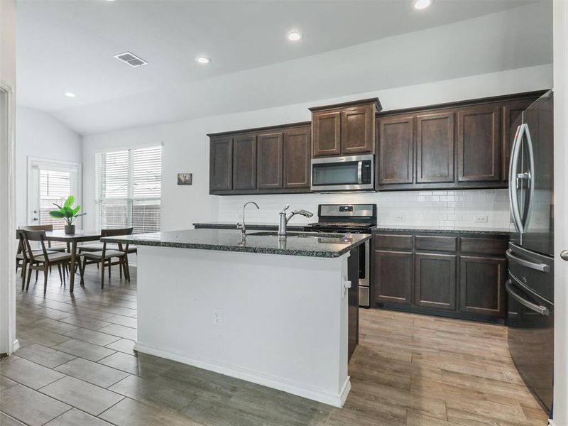 Kitchen featuring light hardwood / wood-style floors, vaulted ceiling, dark stone countertops, a kitchen island with sink, and appliances with stainless steel finishes