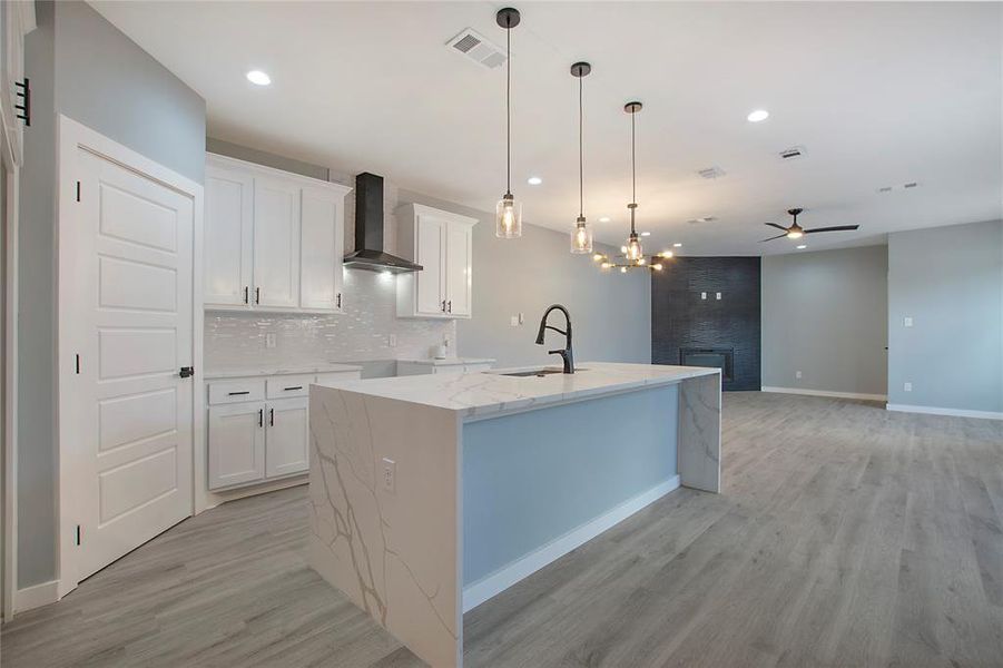 Kitchen with ceiling fan with notable chandelier, wall chimney exhaust hood, a kitchen island with sink, sink, and white cabinets