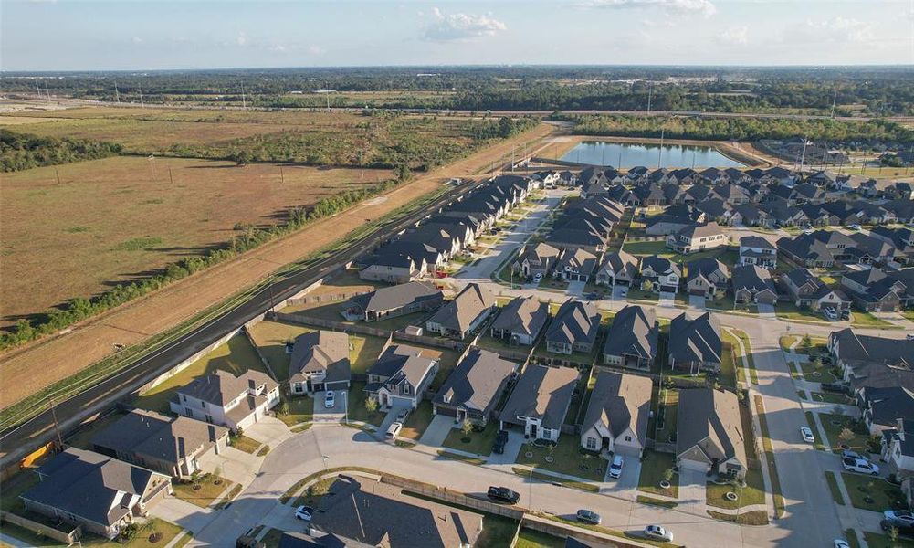 This aerial photo showcases a suburban housing development with uniform, single-family homes, neatly arranged along curving streets. A pond is situated within the community, and there's open land nearby, indicating potential for expansion or natural surroundings. The area is easily accessible via a main road that runs alongside the neighborhood.