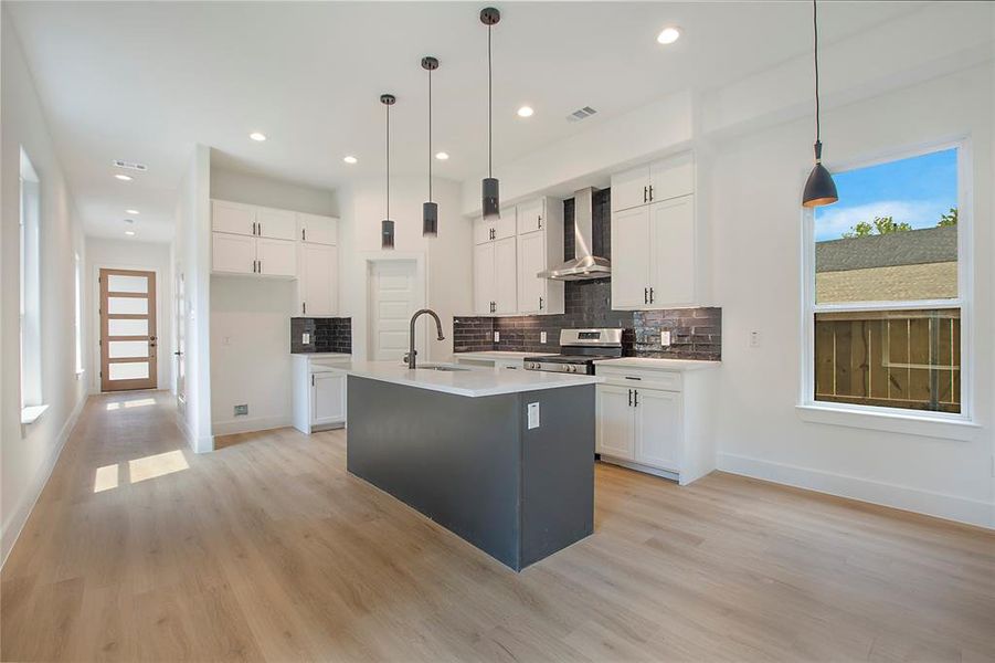 Kitchen featuring wall chimney exhaust hood, a center island with sink, stainless steel stove, sink, and white cabinetry