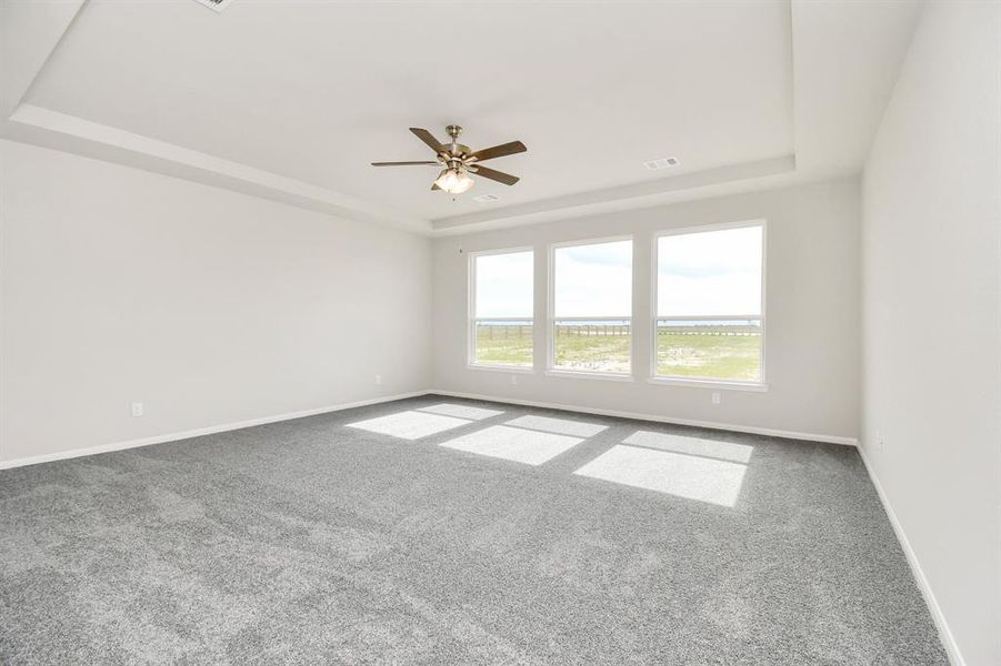 Step into the the primarybedroom with plush carpet, high tray ceiling, ceiling fan, and large windows.