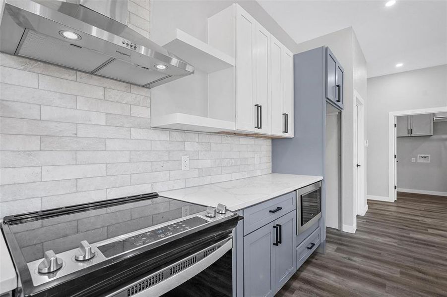 Kitchen featuring dark wood-type flooring, light stone counters, stainless steel microwave, stove, and wall chimney range hood