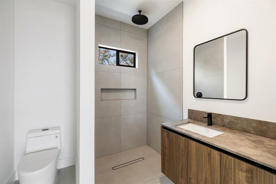 This is a modern bathroom featuring a wall-mounted toilet, a sleek vanity with a wooden finish and a rectangular vessel sink, a large mirror with a black frame, and a high window allowing natural light.