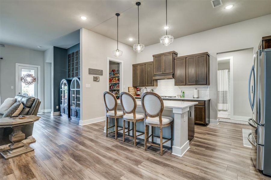 Kitchen with dark brown cabinetry, decorative light fixtures, backsplash, stainless steel refrigerator, and light hardwood / wood-style floors