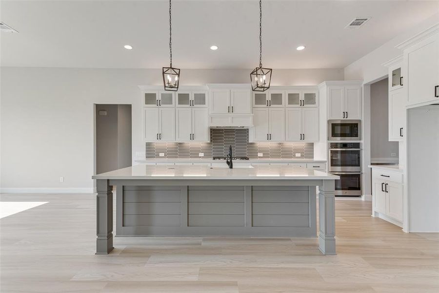 Kitchen featuring tasteful backsplash, white cabinets, hanging light fixtures, a center island with sink, and appliances with stainless steel finishes