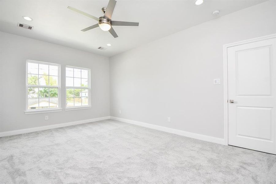 This is a bright, spacious  Primary bedroom with neutral walls and carpet flooring, featuring a ceiling fan and ample natural light from two large windows. A clean, white door adds to the room's modern feel.