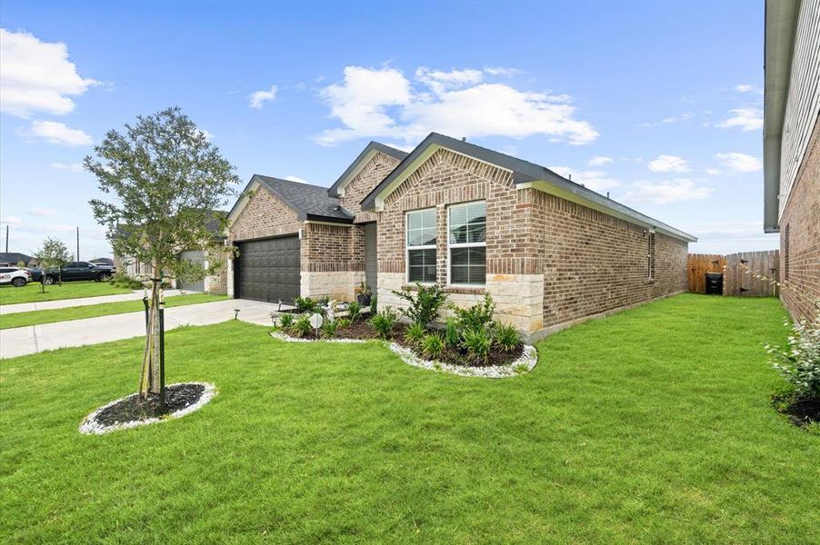 This is a modern single-story brick home featuring a well-manicured lawn, landscaping, and a two-car garage. The property boasts a spacious side yard with a young tree and privacy fencing.