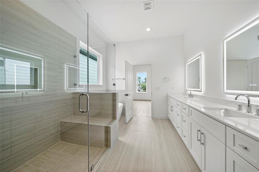 This is a modern, spacious bathroom featuring a large walk-in shower with glass doors, dual vanity sinks with ample storage, and a neutral color palette that enhances the bright, airy feel. Natural light streams in through a window, and high-quality finishes are evident throughout.