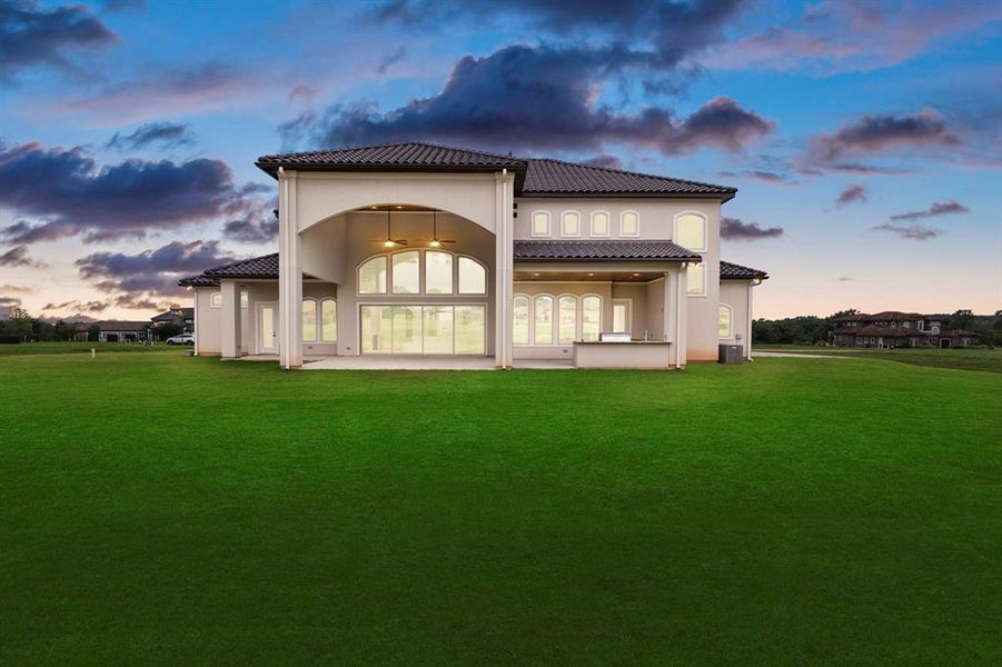 This is a spacious two-story home featuring a modern Mediterranean design with a tiled roof, large arching windows, and an expansive covered patio, set on a generous green lawn.