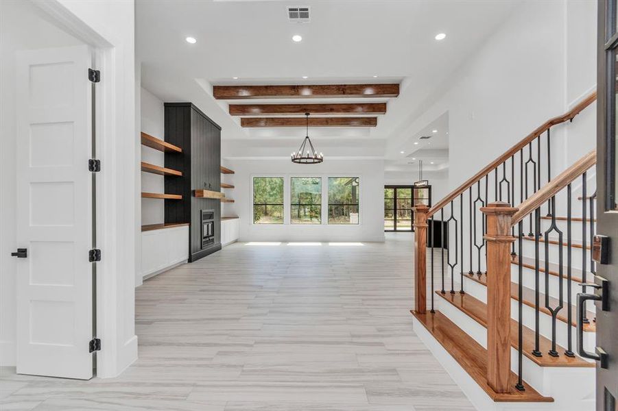 This is a spacious, modern open-plan interior with high ceilings accented with wooden beams, light tile floors, and abundant natural light from large windows. The area features a stylish staircase with wooden handrails and wrought-iron balusters, leading to an upper level.