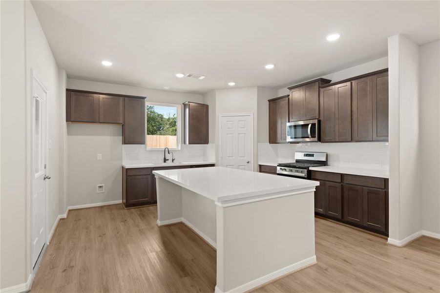 This light and bright kitchen features a large quartz island, dark stained cabinets, a large sink overlooking your family room, recessed lighting, and beautiful backsplash.