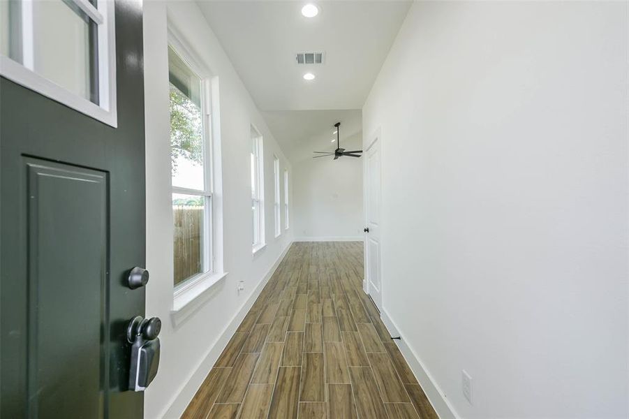 This is a bright and modern hallway featuring wood flooring, multiple windows providing natural light, a fresh white wall color, a ceiling fan, and a contrasting dark entry door.